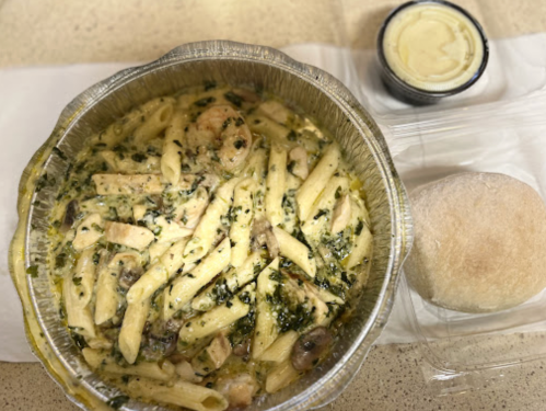 A foil container with creamy pasta, chicken, and spinach, accompanied by a roll and a small container of sauce.